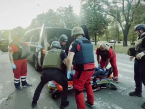 Працівники ДСНС та медики у бронежилетах транспортують пораненого на ношах до автомобіля після російських обстрілів на Нікопольщині