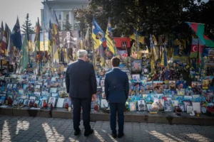 Двое мужчин в костюмах стоят у мемориала с фотографиями военных и флагами