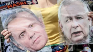 Жінка в жовтому топі тримає дві маски з обличчями Туска і Качинського