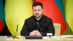 Владимир Зеленский сидит за столом с микрофоном на фоне флагов